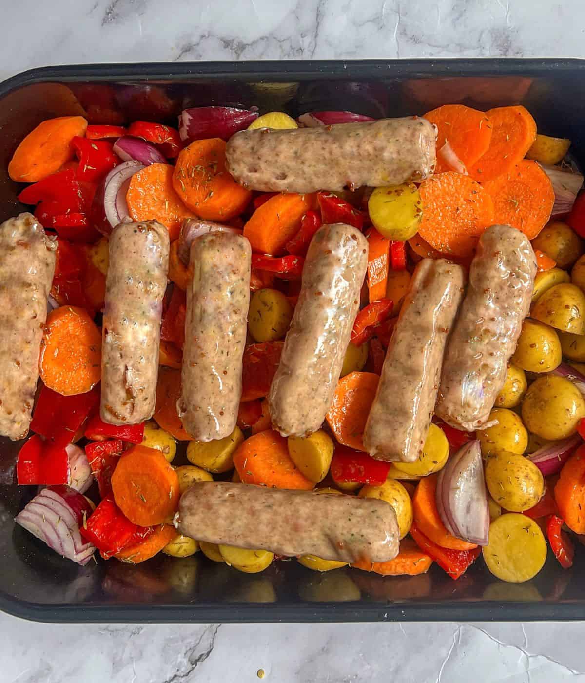 Raw sausages sitting on top of potatoes, carrot, red onions and peppers in a roasting tin.