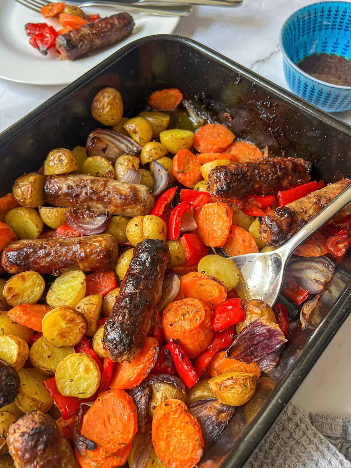Sausages, potatoes and vegetables in a roasting tray with a large spoon in it.