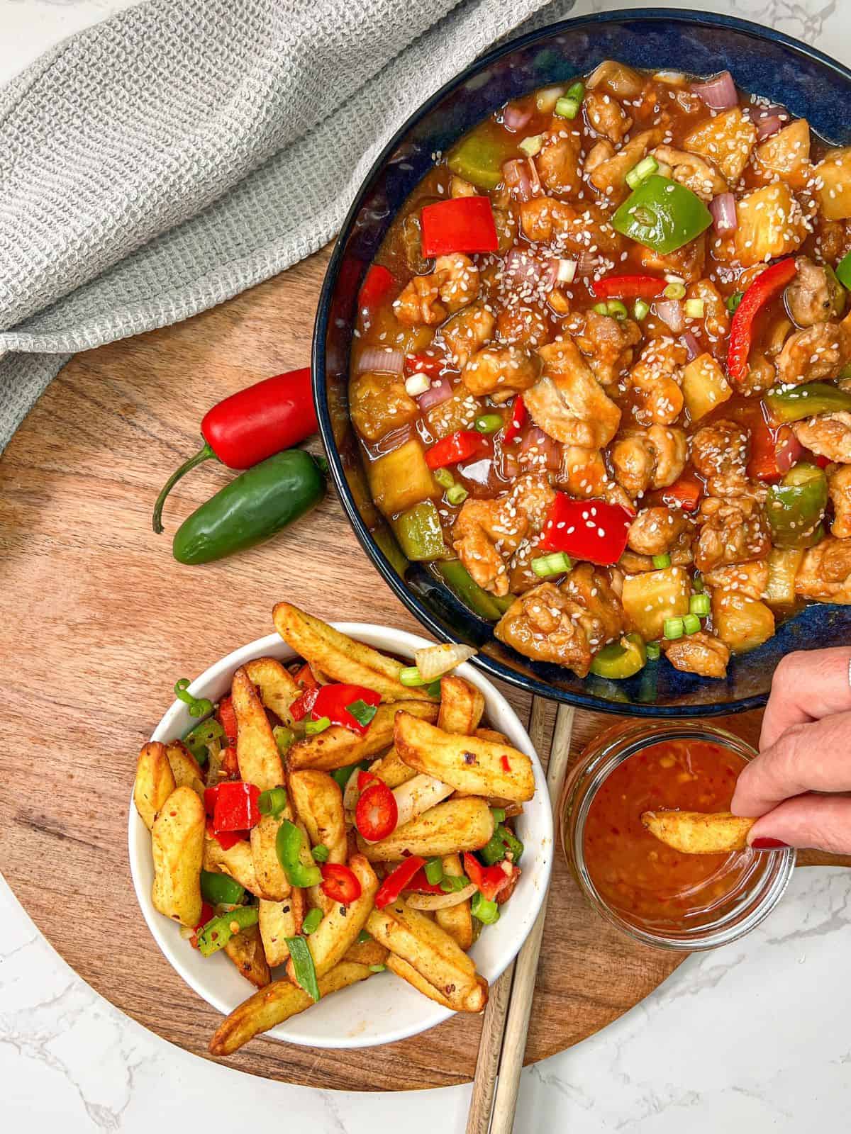 Sweet & sour chicken and salt and pepper chips in bowls, with a bowl of sweet chilli sauce to the side, with a chip being dipped into it.