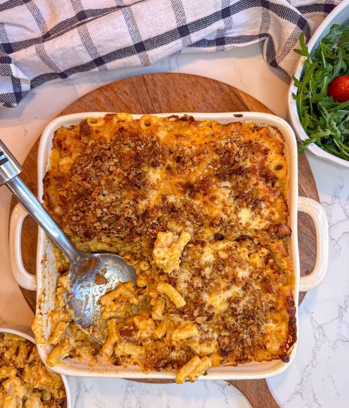 A baking dish containing mac and cheese, with a serving spoon in it. Rocket salad to the side.