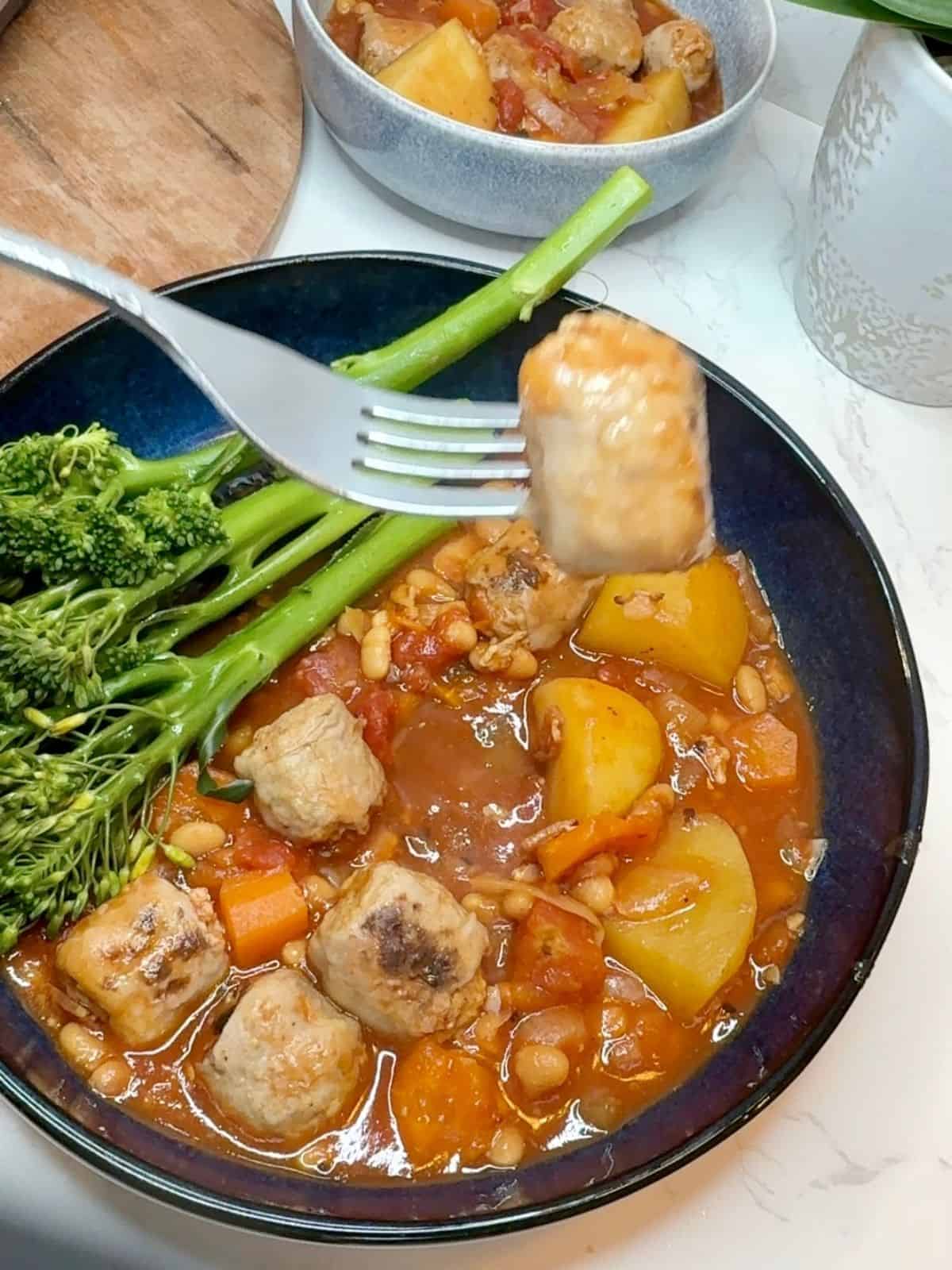 Someone sticking a fork into a bowl of sausage casserole and tenderstem broccoli.