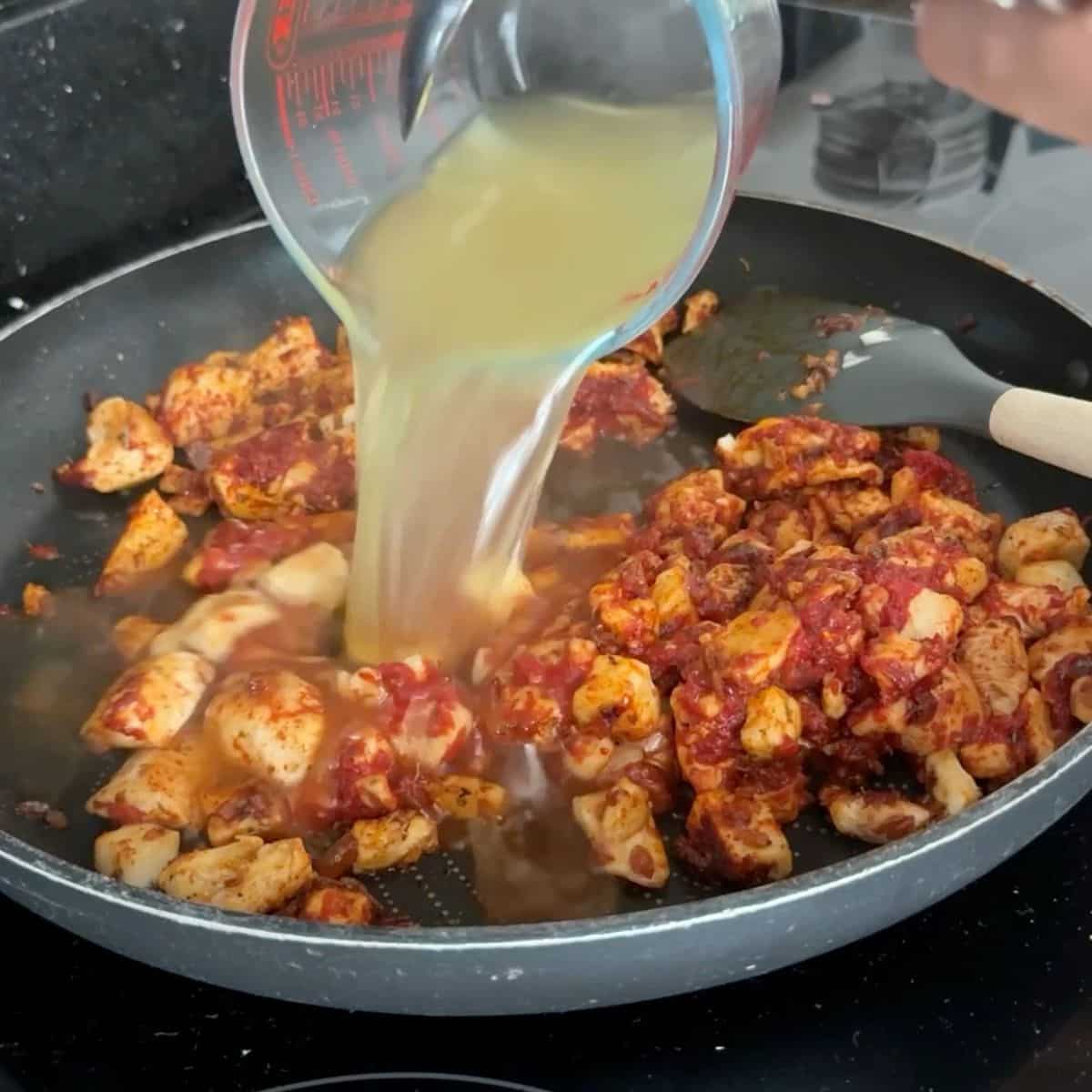 Stock being poured into a frying pan containing halloumi in tomato puree.