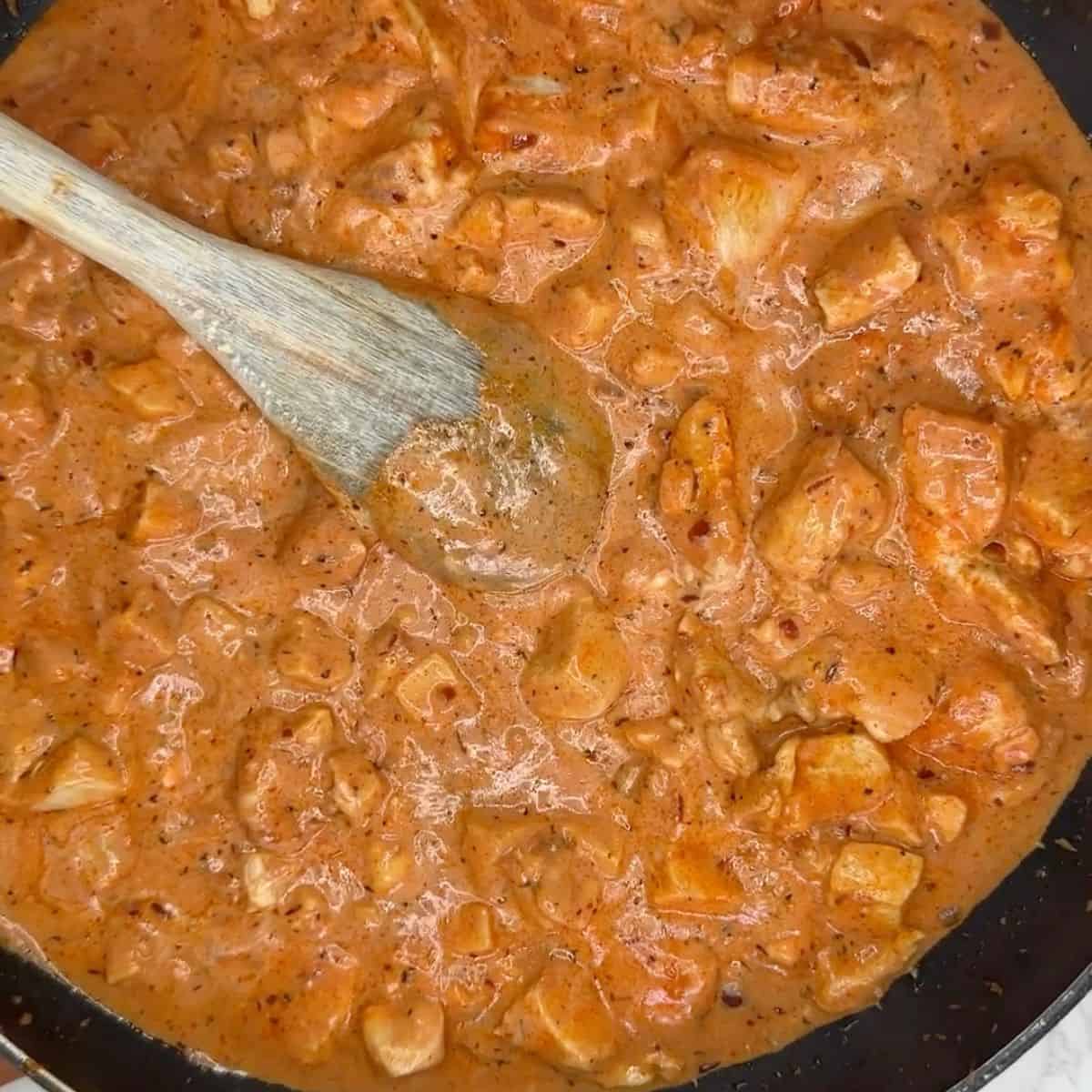 chicken in a creamy tomato sauce in a frying pan, with a wooden spoon in the sauce.