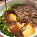A plate of beef brisket, roast potatoes & broccoli with gravy being poured over it.
