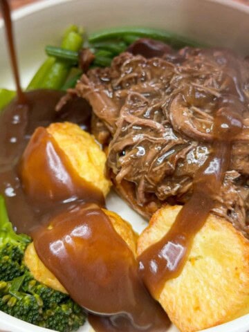 A plate of beef brisket, roast potatoes & broccoli with gravy being poured over it.
