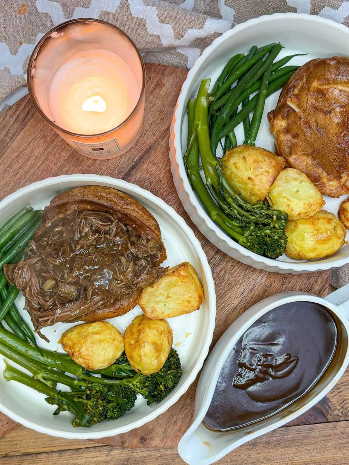 Two plates containing yorkshire puddings, beef brisket, roast potatoes & broccoli with a jug of gravy and a candle on the side.