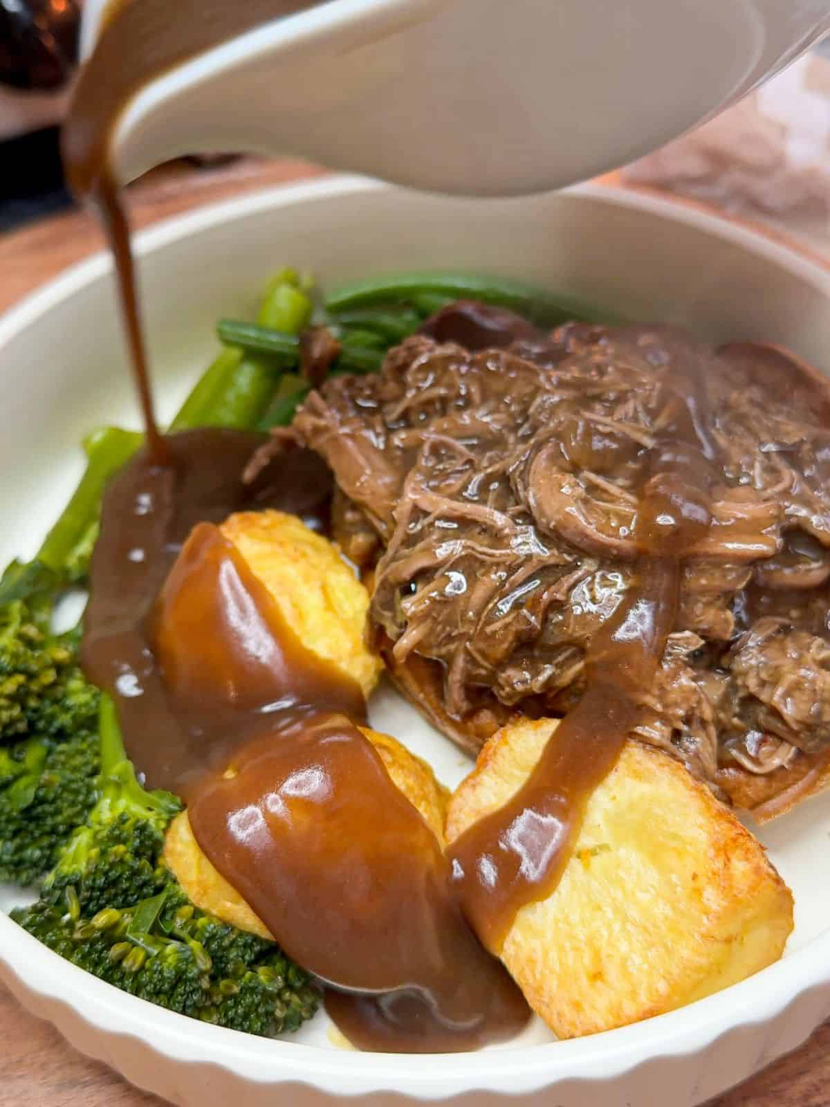 A plate of beef brisket, roast potatoes & broccoli with gravy being poured over it.