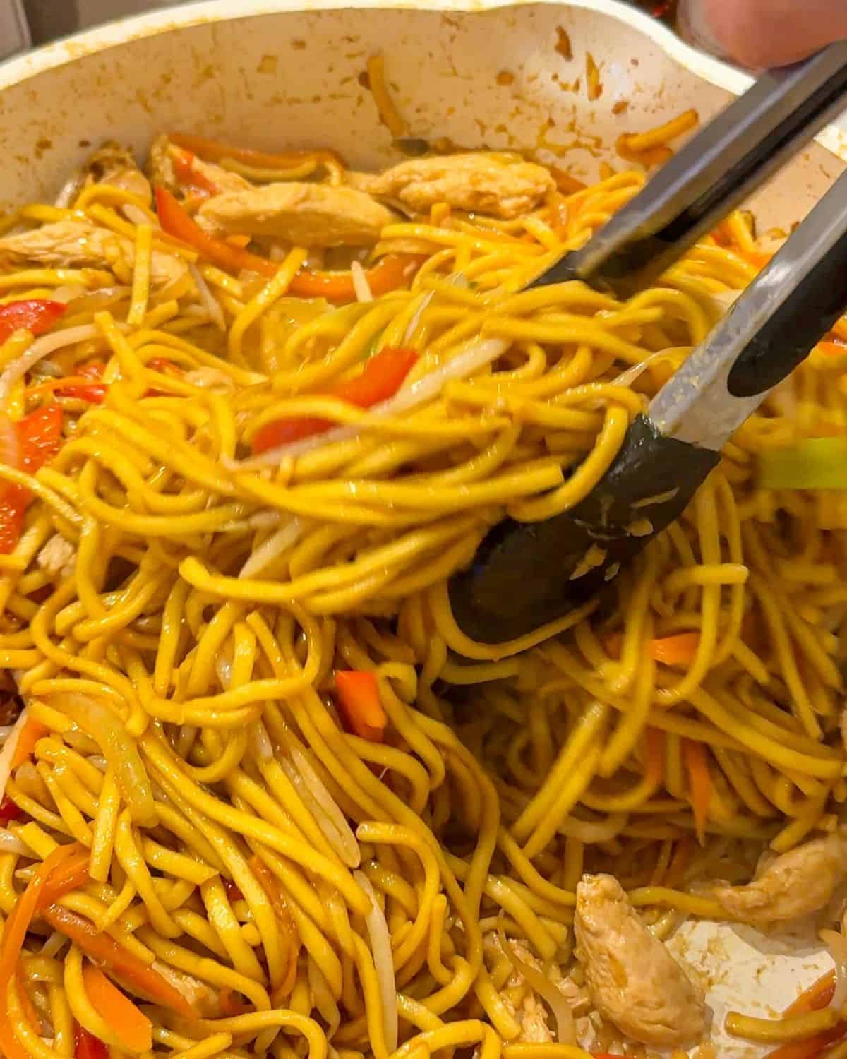 Chicken chow mein being stirred with tongs in a frying pan.