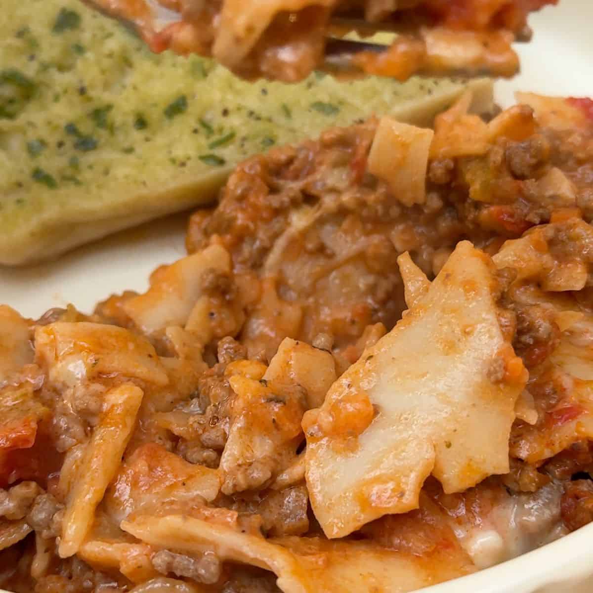 A plate of one pan lasagne with garlic bread in the background.