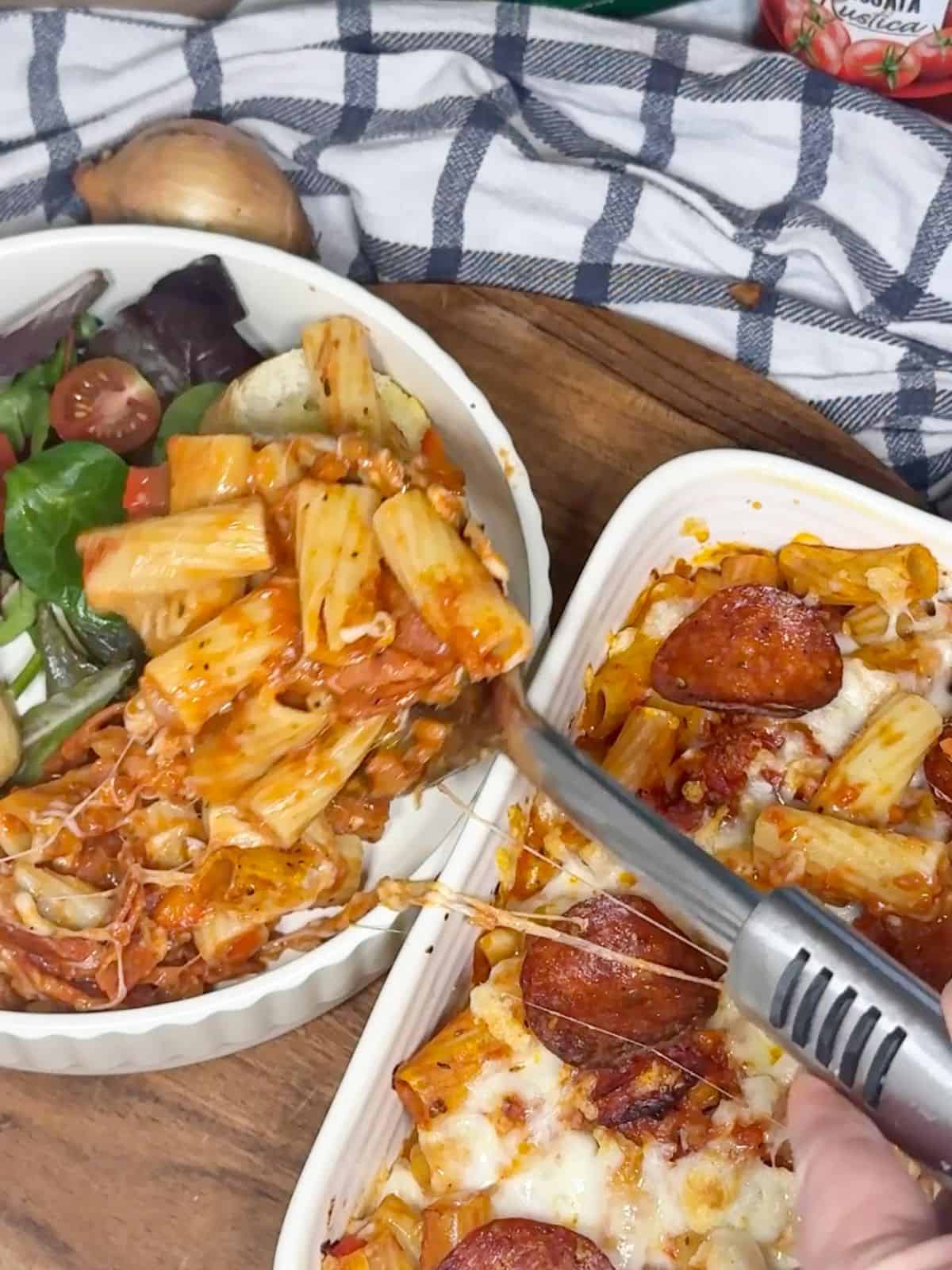 A large dish of pepperoni pizza pasta, with a bowl of the pasta with salad next to it.