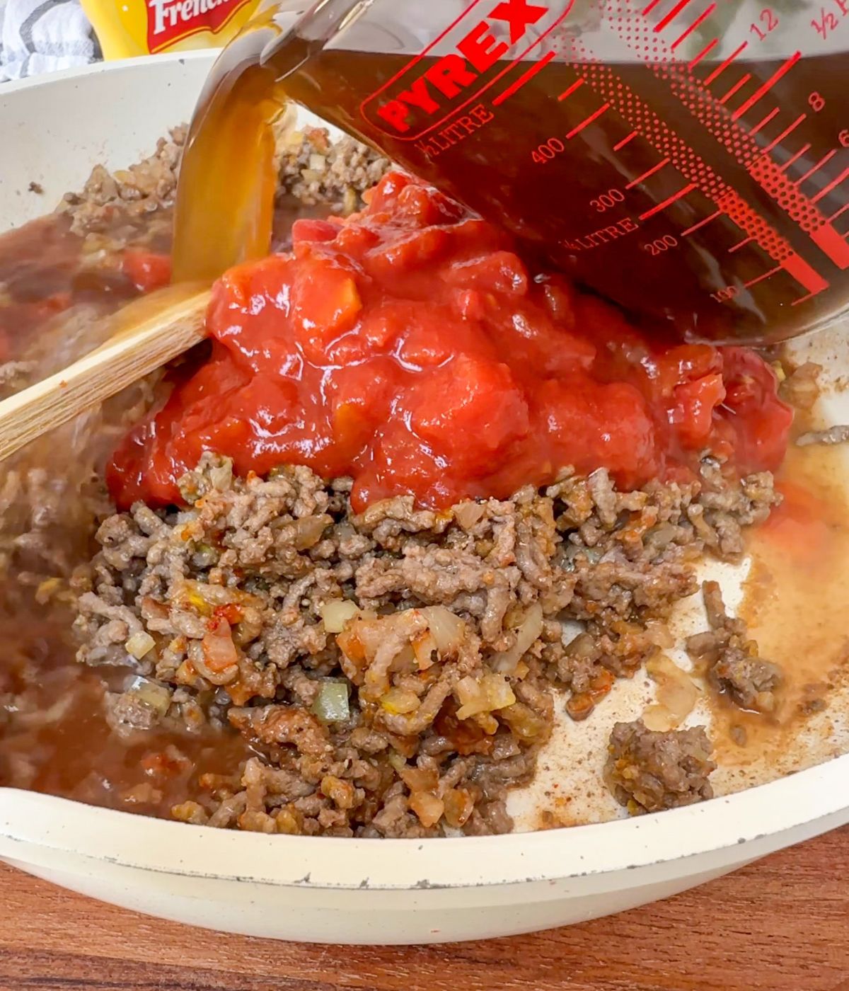Beef stock being poured into a pan containing chopped tomatoes and mince.
